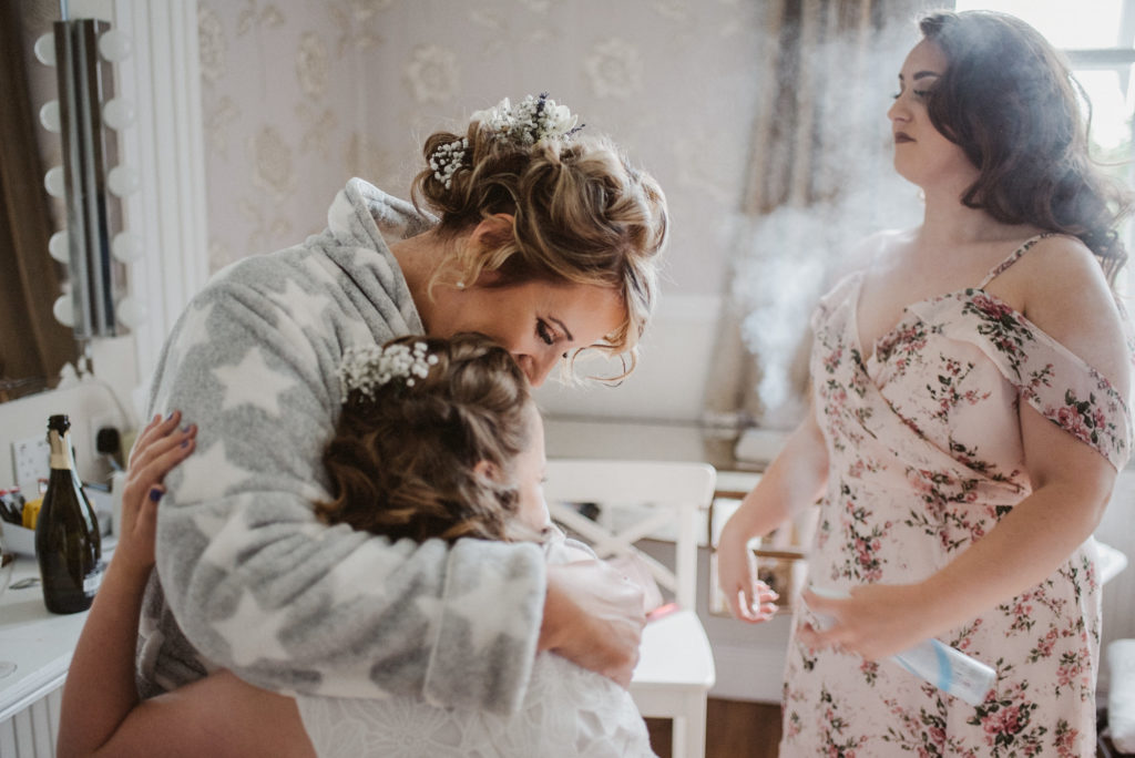 Bride hugging a young girl whilst a woman sprays perfume in the background