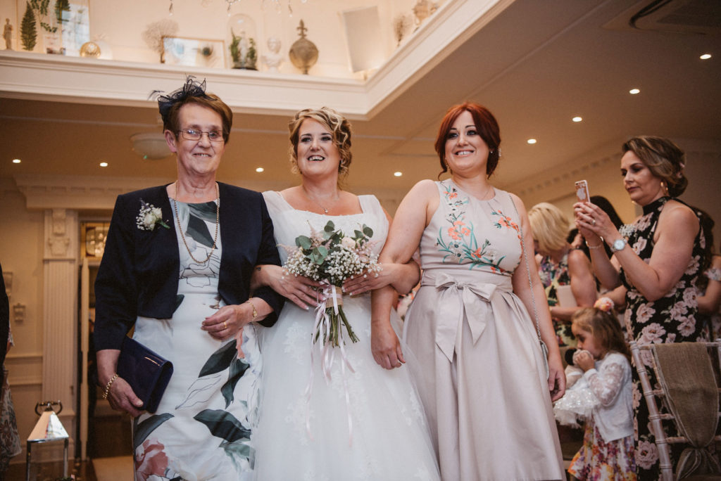 Bride walks up the aisle smiling with her mother and daughter in the langtree suite