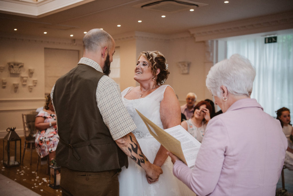 Bride looks happily at groom holding hands before the celebrant in the langtree suite