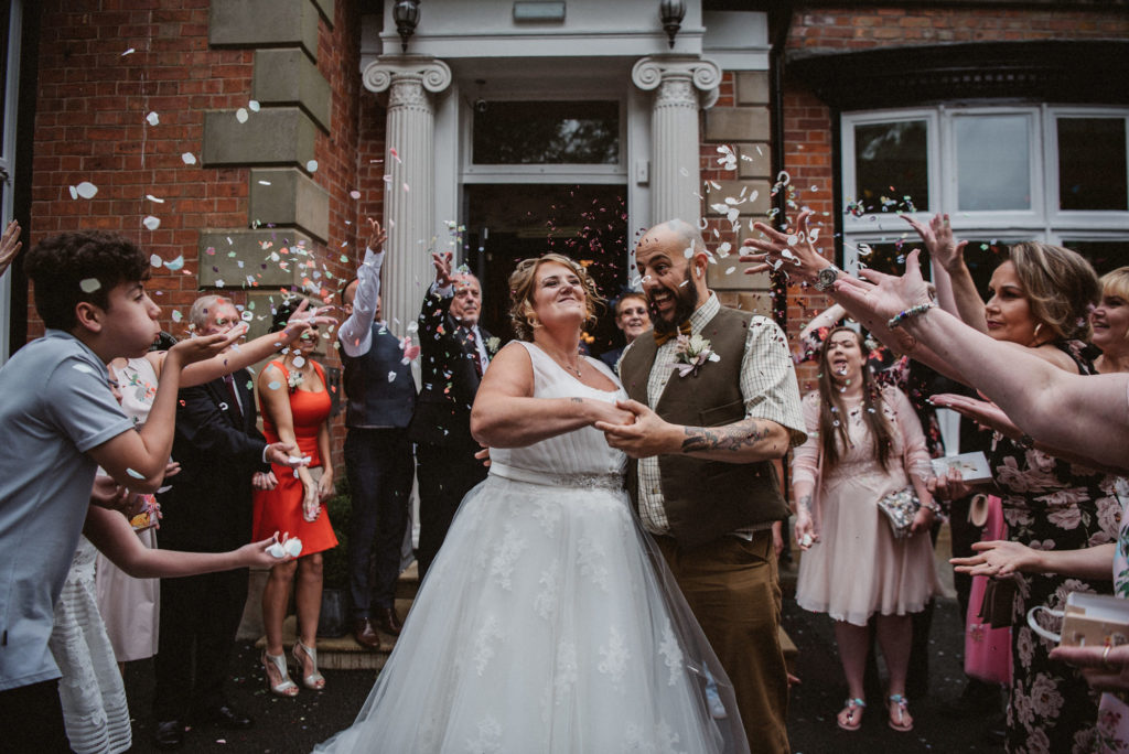 Bride and groom react to confetti being thrown at them on the front steps of Ashfield House