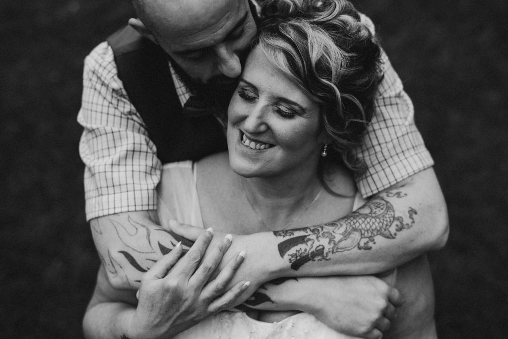 Groom stands behind bride and kisses her on the cheek in a romantic embrace