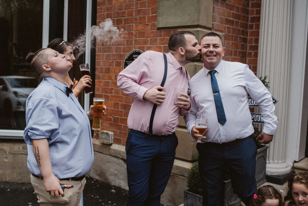 A man wearing a pink shirt kisses a man wearing a white shirt on the cheek whilst a lady blows steam from her e cigarette into the air during a wedding