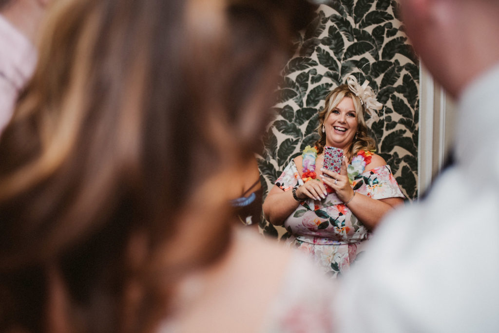 A wedding guest wearing a necklace made from flowers laughs at two other wedding guests whilst holding her phone