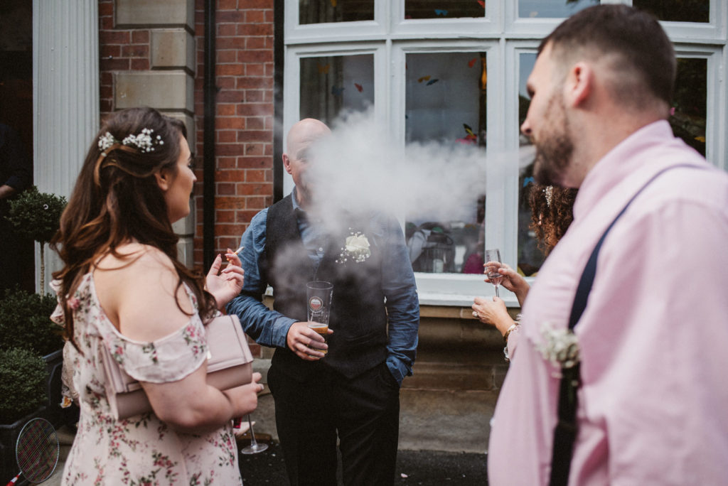A wedding guest blows smoke from his e cigarette into the face of another wedding guest