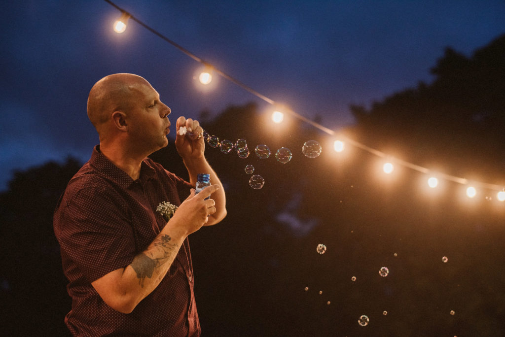 Wedding guest blows a chain of bubbles whilst standing next to a string of fairy lights