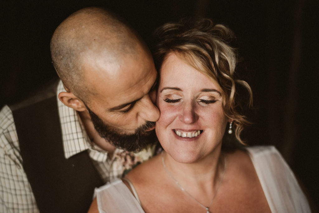 The groom kisses the bride on the cheek in a romantic portrait in a small patch of light at golden hour