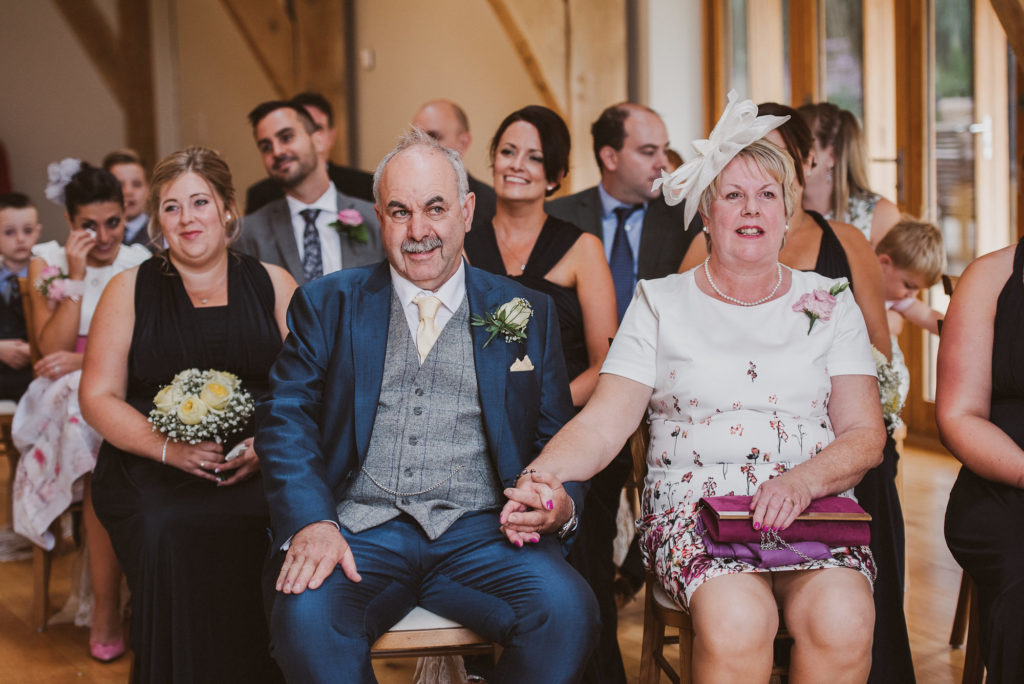 mother of the bride and mother of the groom look happily at the bride and groom during their barn ceremony