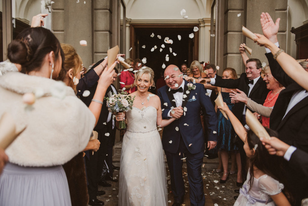 bride and groom get bombarded with confetti thrown by their enthusiastic wedding guests