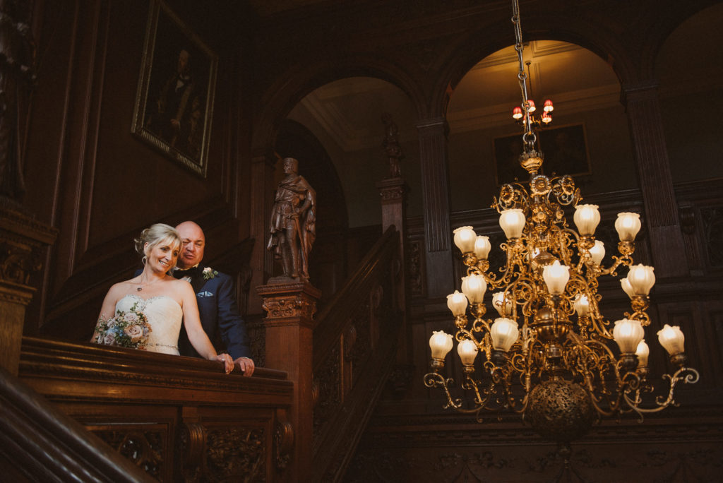 bride and groom look down the stairs next to a big chandelier