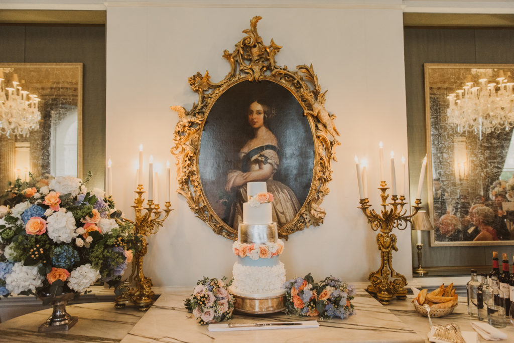 a four tier silver and white wedding cake with pink roses sits under a grand painting in the French Dining Room
