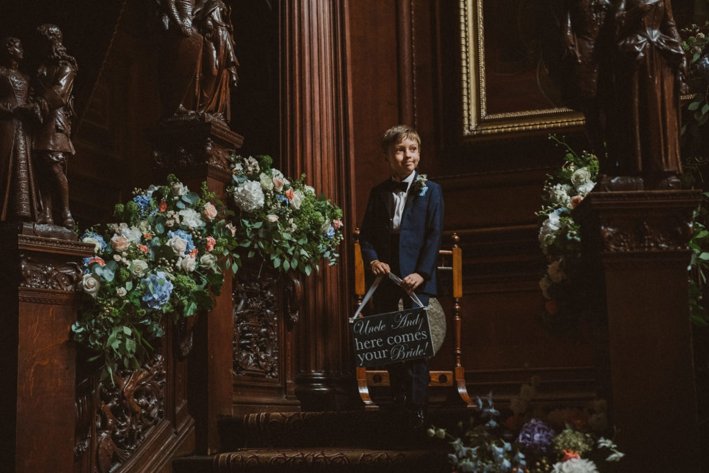 page boy holding sign walks down aisle ahead of bride during the wedding ceremony in the Great Hall