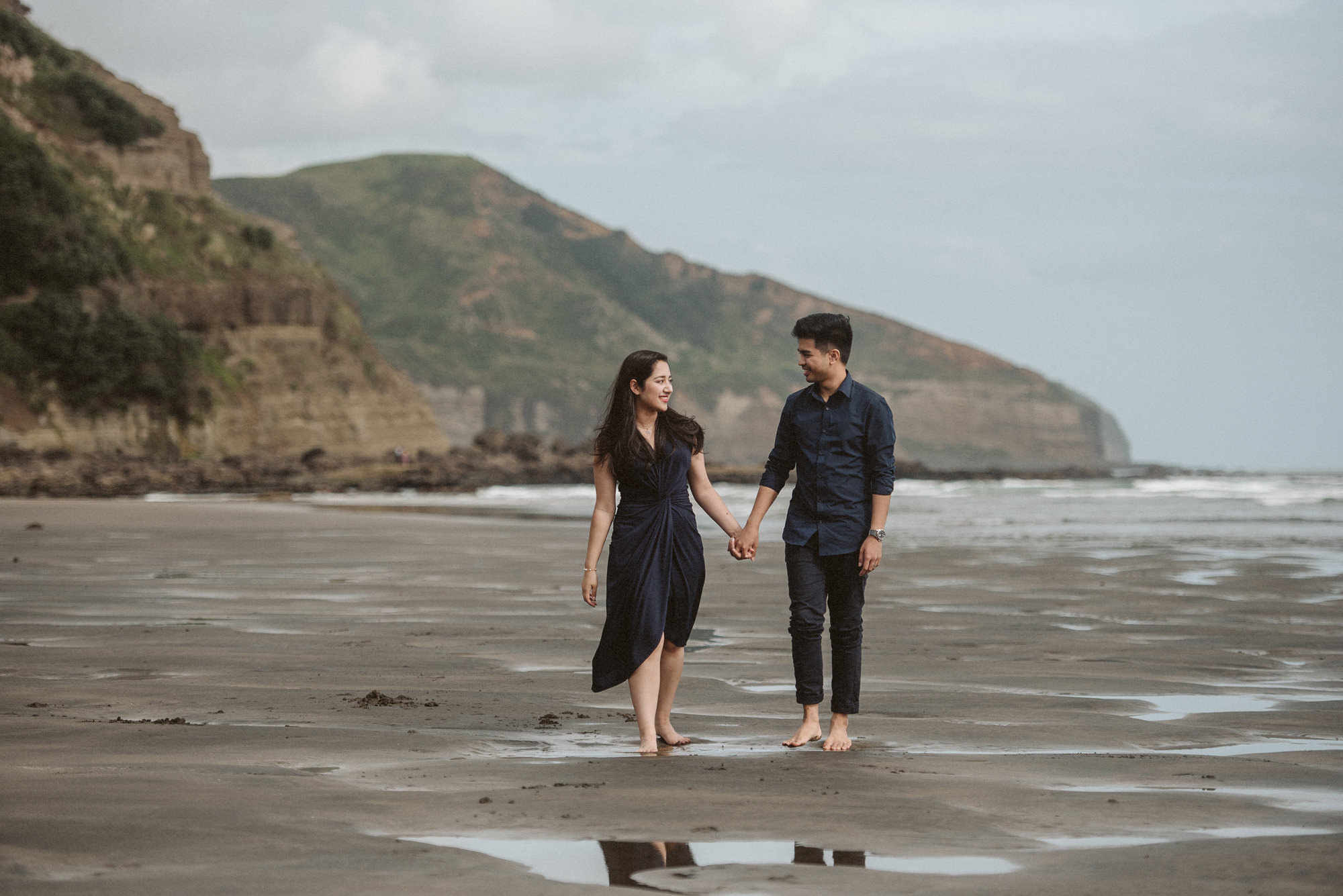 A couple stroll along the beach for their engagement photo session at Maori Bay in Auckland
