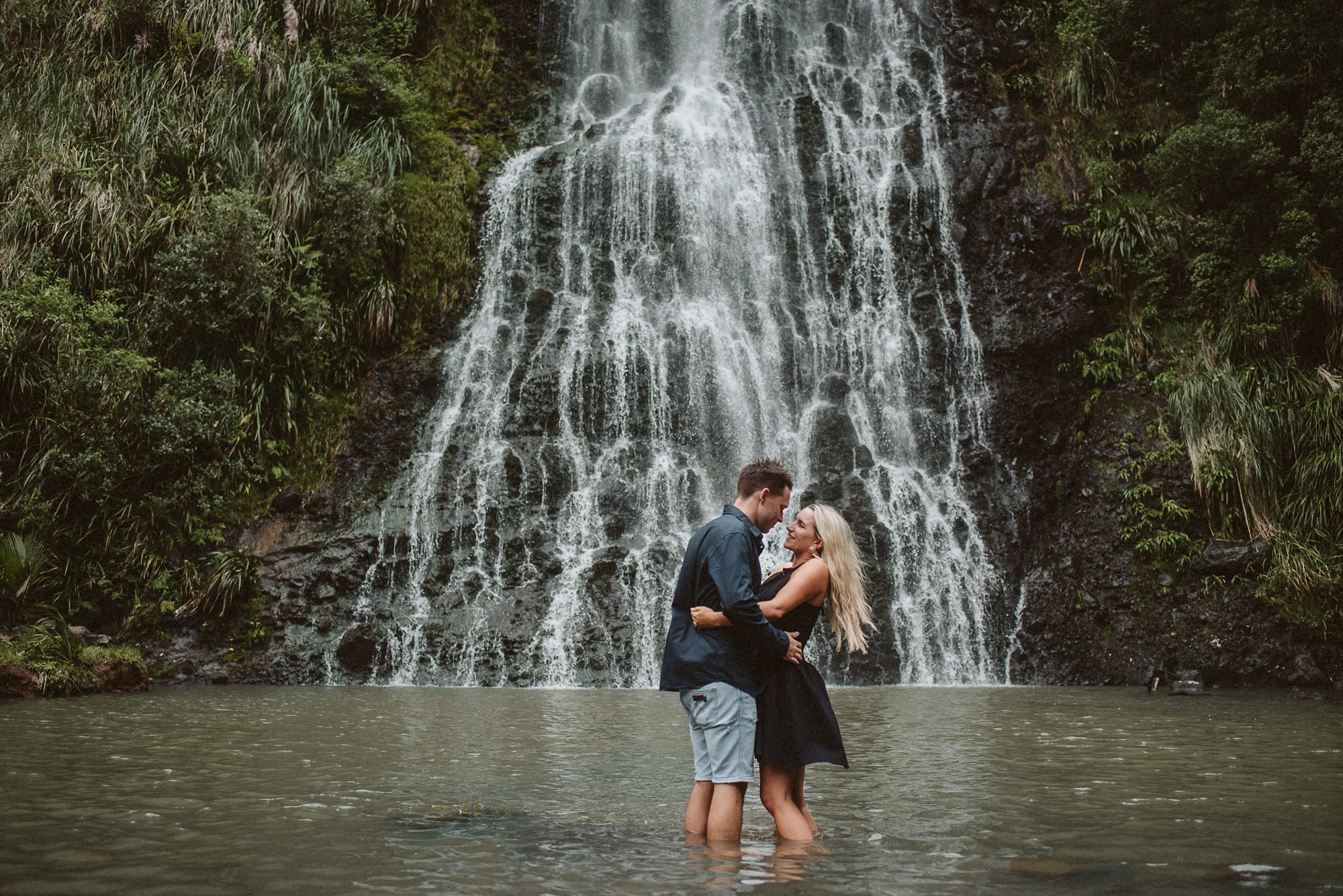 An engaged couple embrace under a waterfall at Karekare Falls, Auckland during their engagement shoot