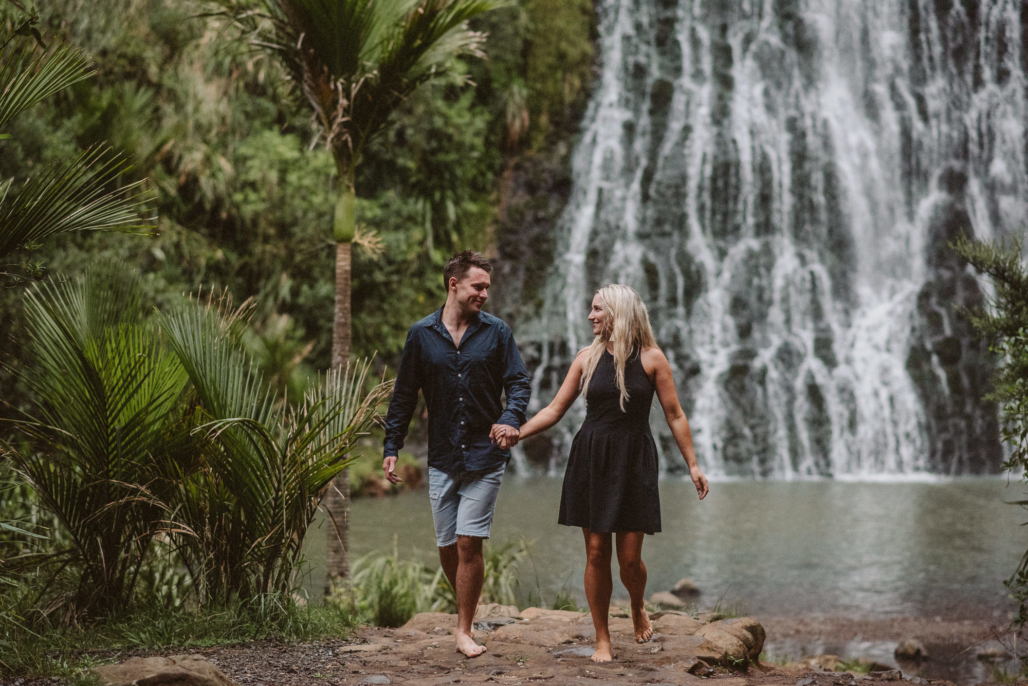 An engaged couple walk along looking at each other happily in front of Karekare Falls in New Zealand