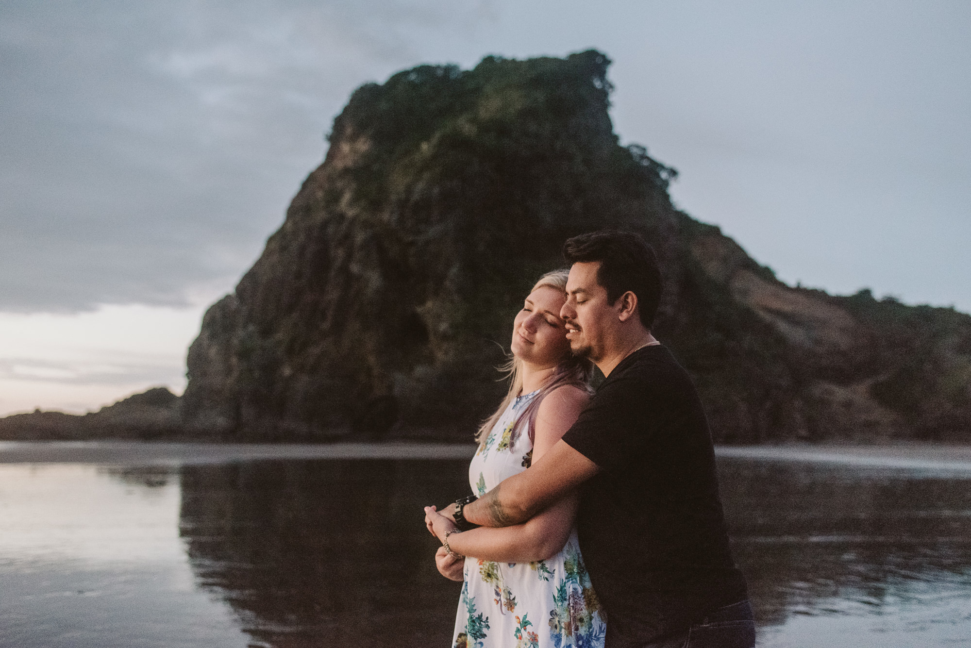An engaged couple have a hug in front of a large rock formation on Piha Beach in Auckland