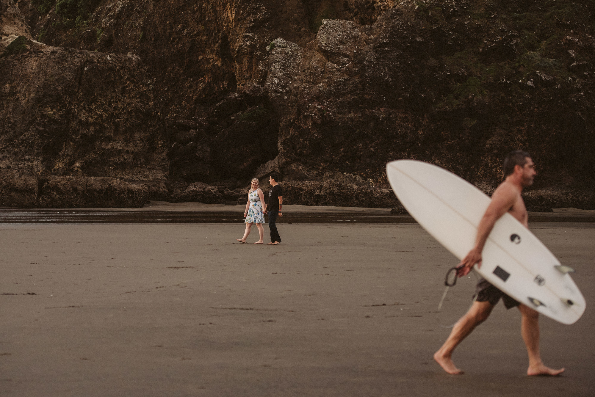 A couple walk along Piha Beach as a surfer walks past
