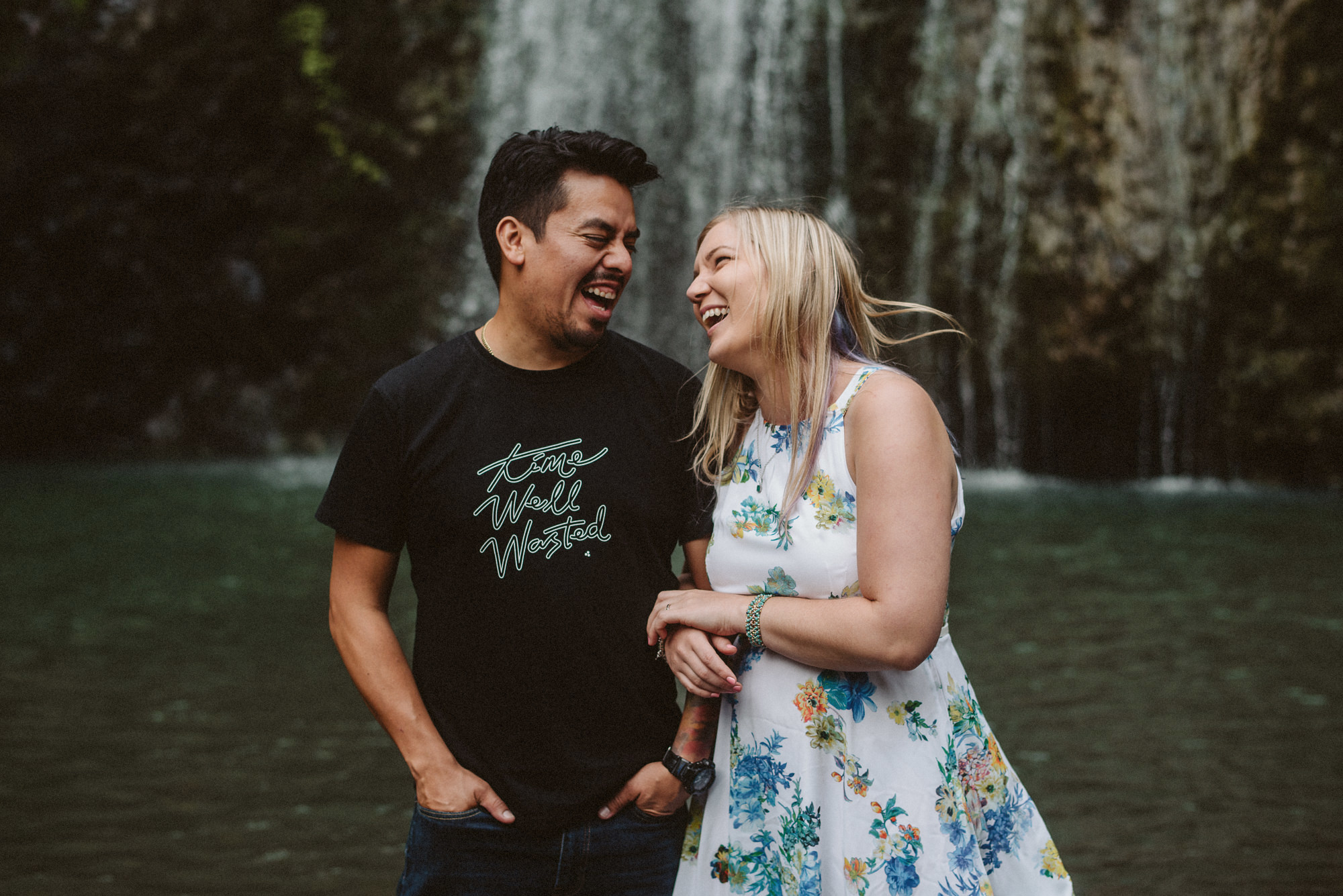 A couple look at each other and laugh in front of Kitekite Falls in Auckland, New Zealand