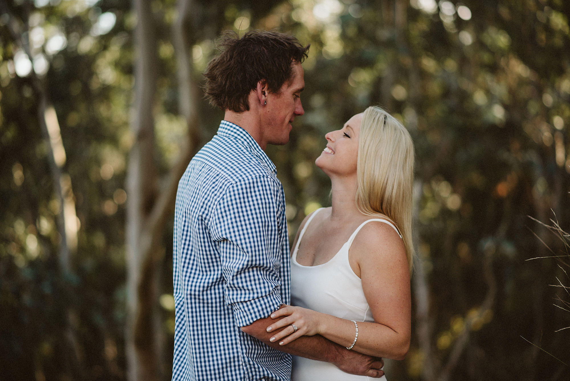 An engaged couple look into each other's eyes in front of some trees