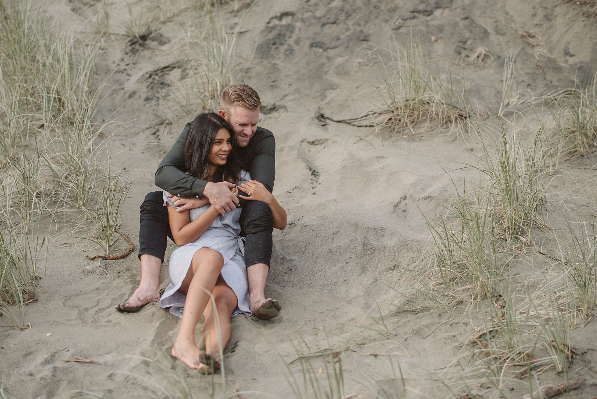 A man embraces a woman sitting on a sand dune at Bethells Beach