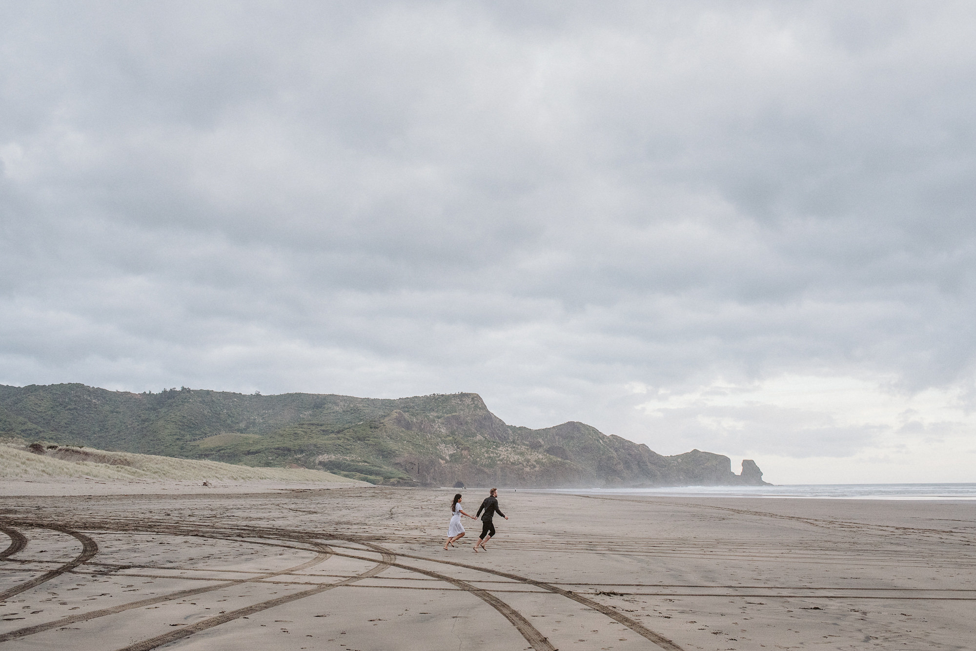 A couple run across Bethells Beach towards the ocean in Auckland, New Zealand