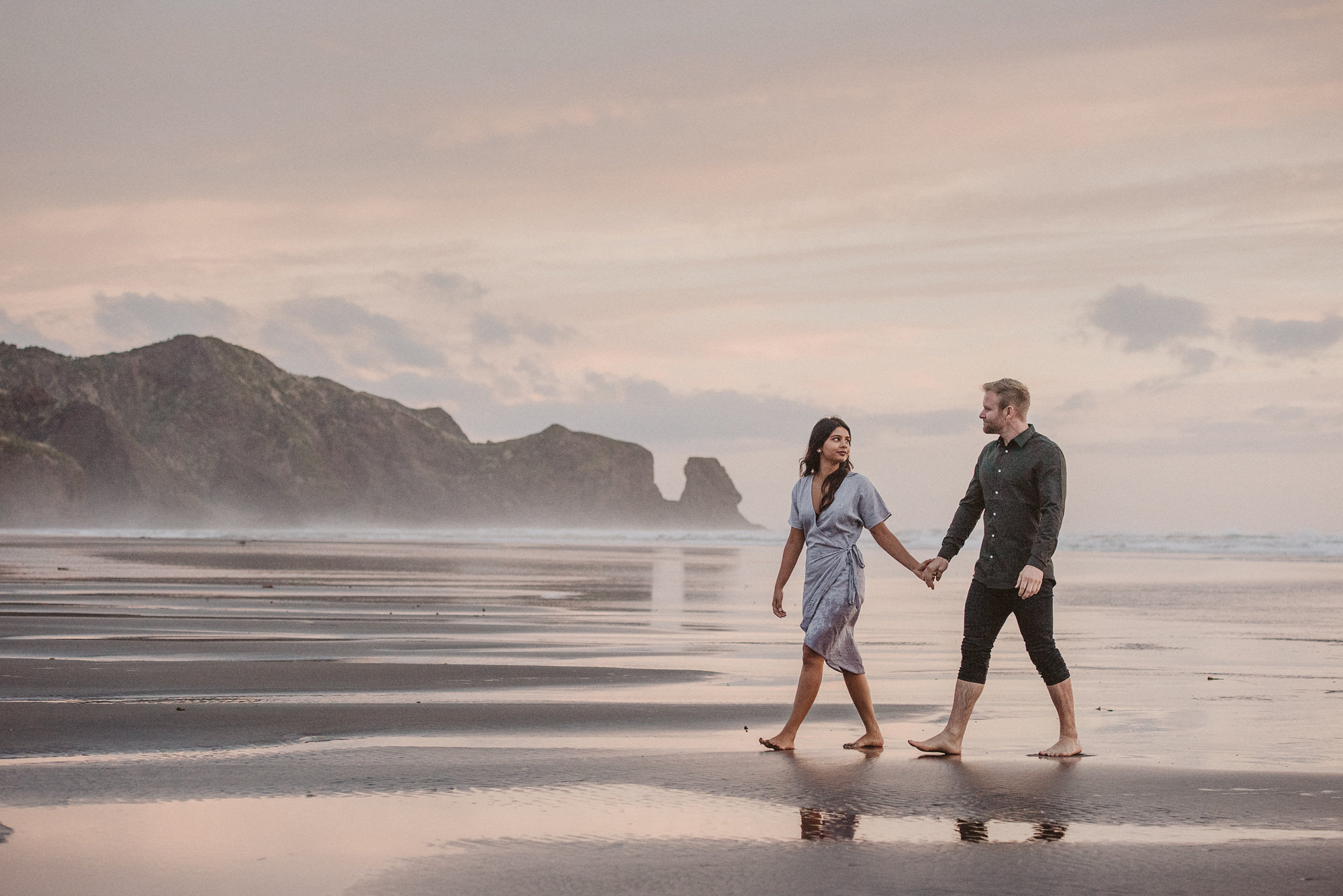 An engaged couple walk along Bethells Beach at sunset