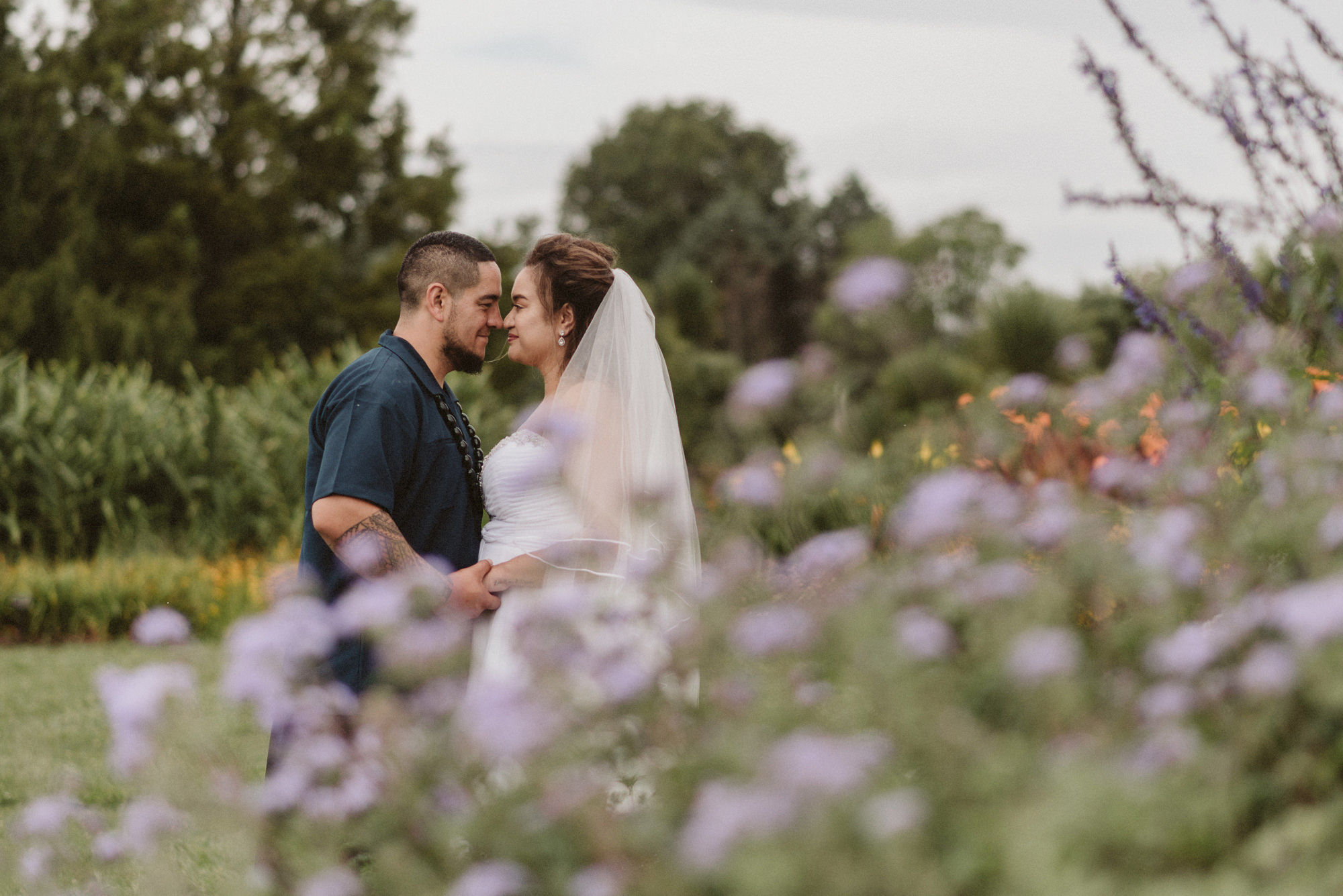 A couple romantically embrace behind some purple flowers at Auckland Botanic Gardens in Manukau, Auckland