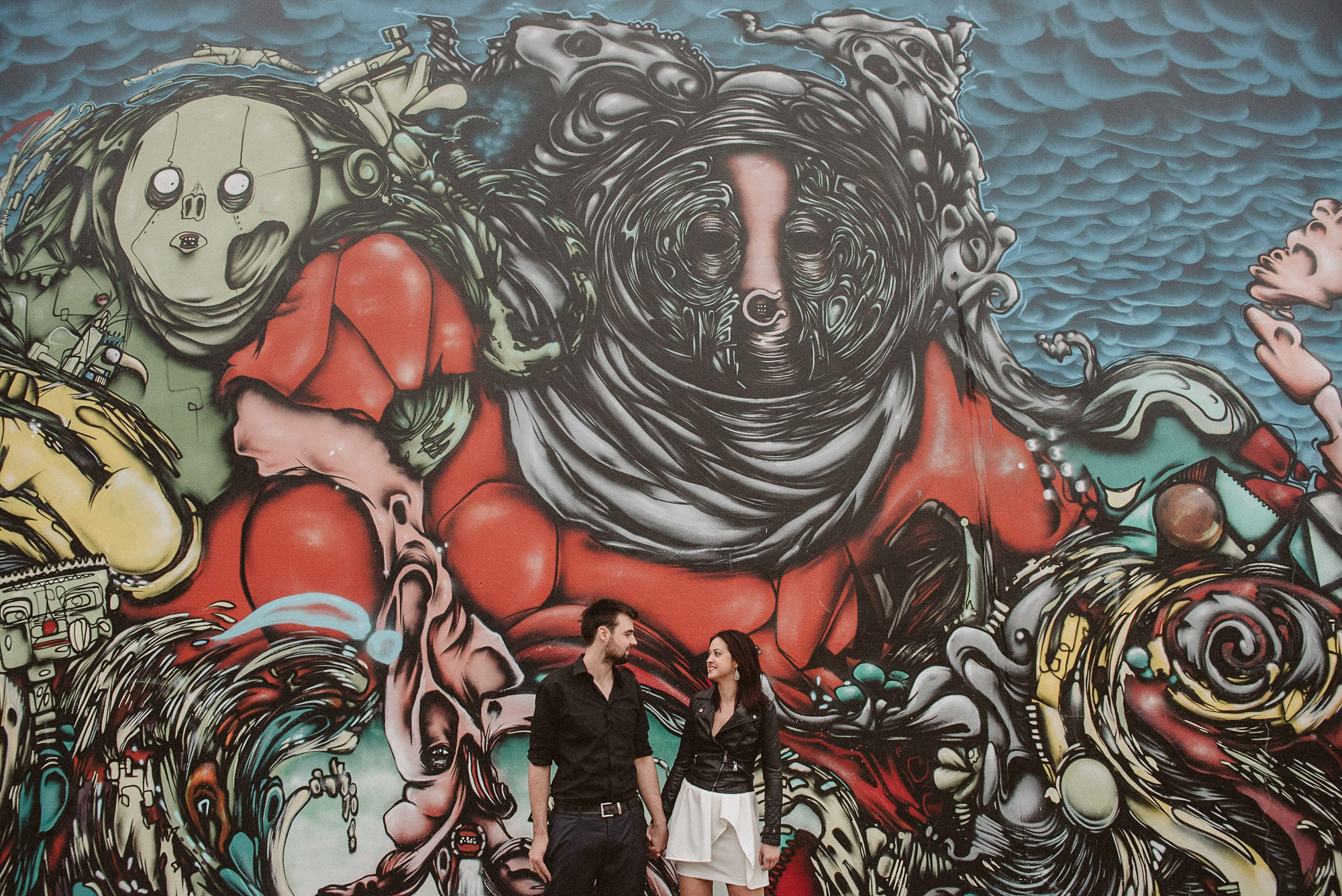 An engaged couple stand in front of a wall painted with street art