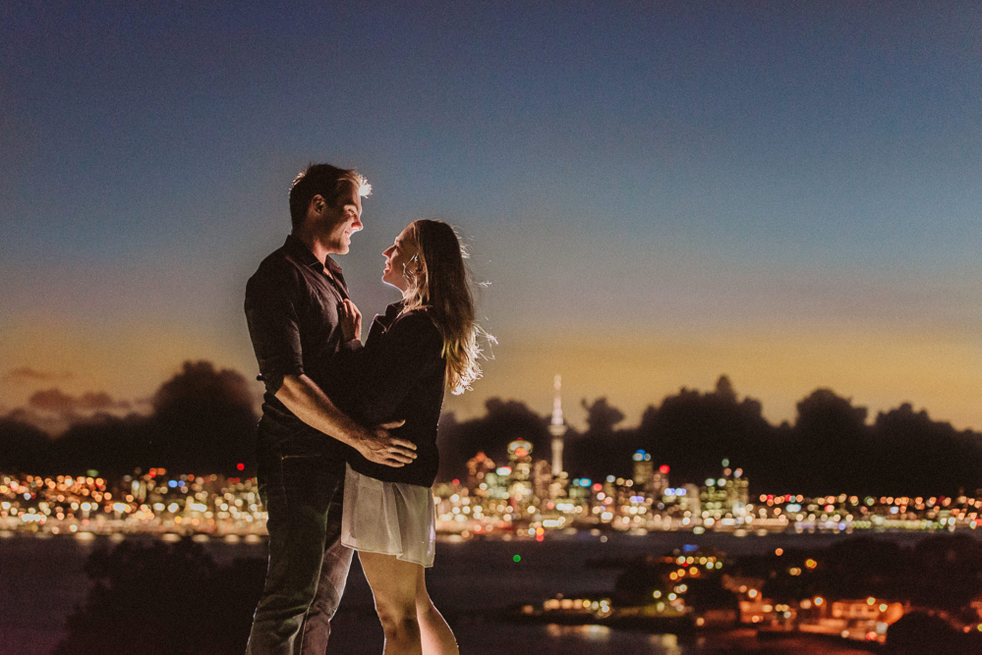 An engaged couple embrace on top of Mount Victoria at night with the central Auckland skyline in the background