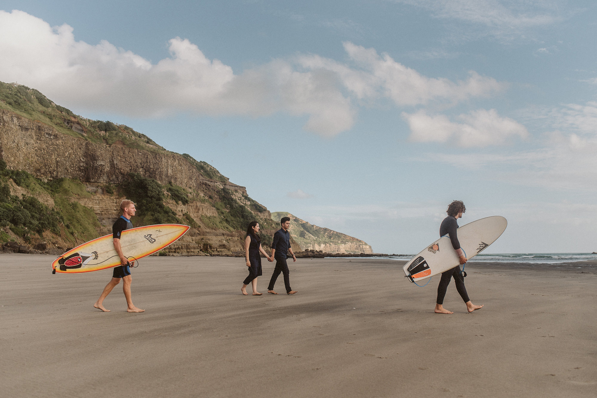 An engaged couple stroll across Maori Bay in Auckland holding hands whilst two surfers walk past